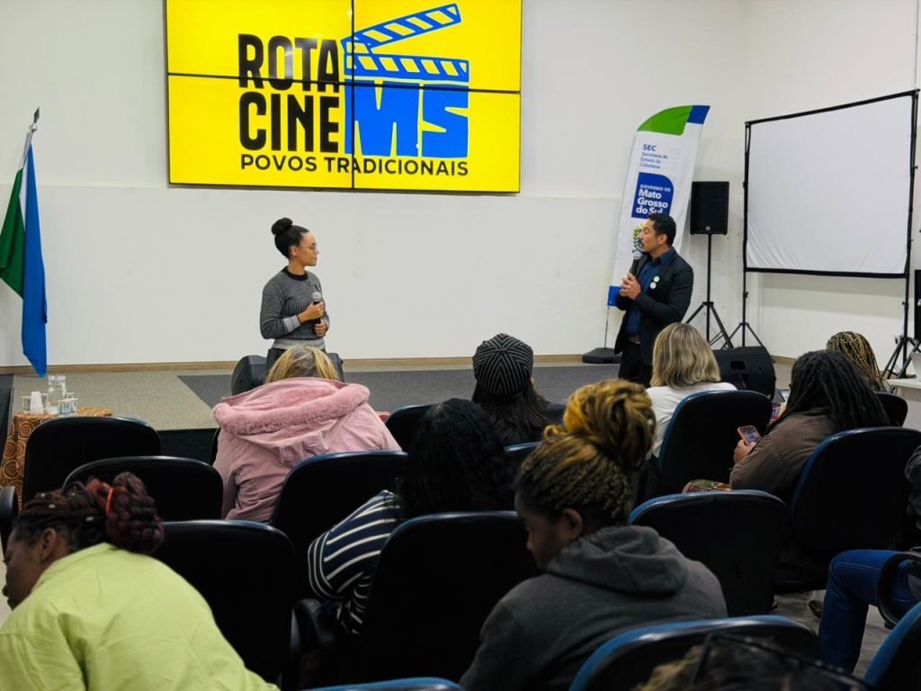 Uma mulher e um homem conversam no palco de um auditório sob um telão que exibe o logotipo "ROTA CINE MS - Povos Tradicionais".