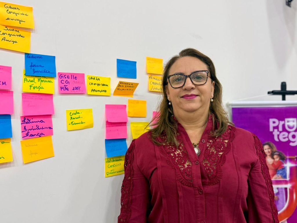 Foto em plano médio e focado de uma mulher de pele clara, cabelos castanhos na altura dos ombros e óculos de grau com armação escura e estampa de tartaruga. Ela sorri discretamente para a foto, vestindo uma camisa de botão na cor vinho com detalhes em renda texturizada no busto e nas mangas. À esquerda, ao fundo, há uma parede branca repleta de post-its coloridos (amarelos, rosas e azuis) com nomes e palavras escritas à mão. À direita, aparece uma parte de um banner roxo com a palavra "Protege".