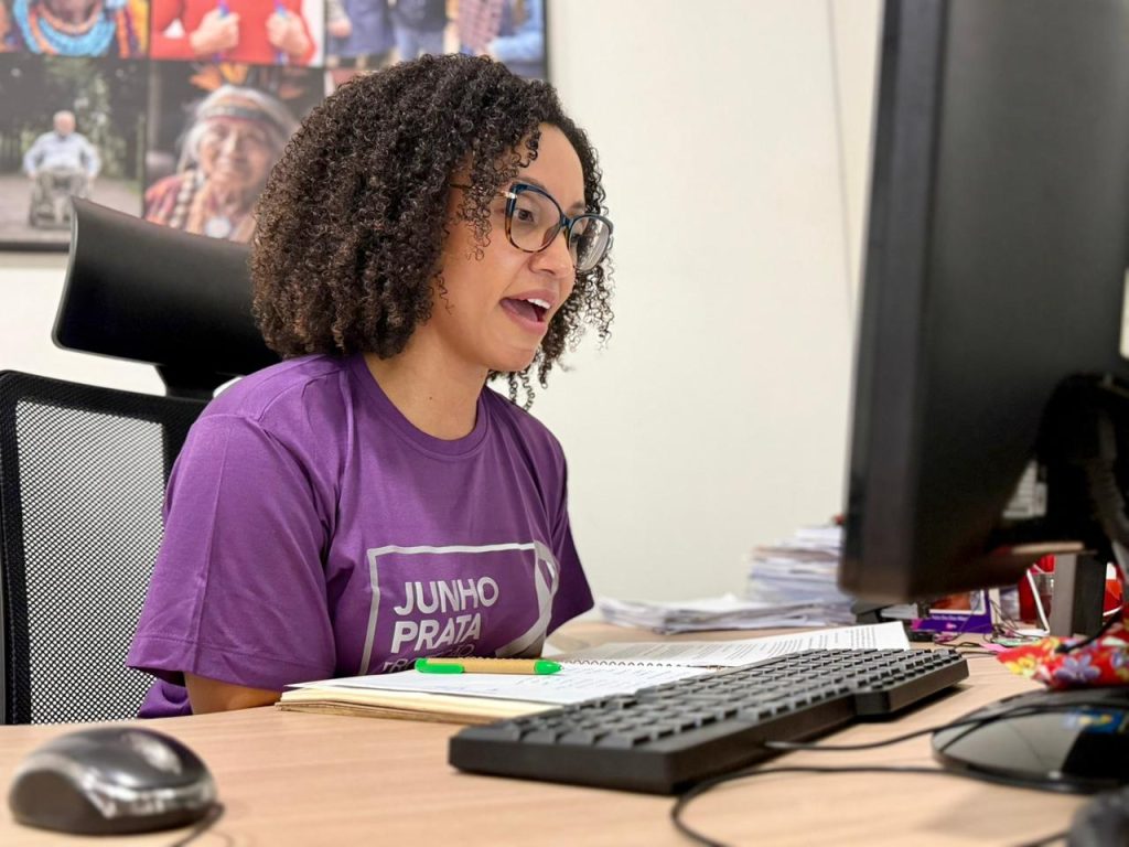 Fotografia colorida em plano médio de uma mulher jovem, com cabelos cacheados e óculos de grau, sentada em uma mesa de escritório. Ela veste uma camiseta roxa com a estampa "Junho Prata" e está de perfil, olhando para o monitor de um computador à sua direita com a boca levemente aberta, indicando que está falando. Na mesa de madeira clara, veem-se um teclado, um mouse e alguns papéis. Ao fundo, uma parede branca com um mural de fotos desfocado.