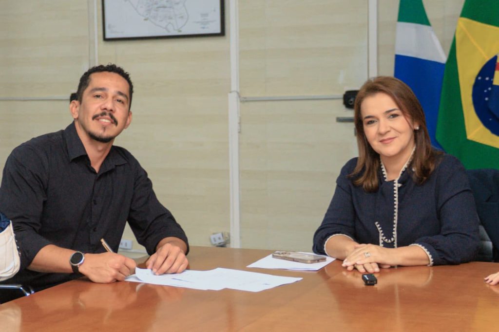 Fotografia em plano médio de duas pessoas sentadas lado a lado atrás de uma mesa de madeira clara. À esquerda, um homem de pele parda, cabelo curto, barba e bigode, vestindo camisa preta, assina um documento com uma caneta. À direita, uma mulher de pele clara, cabelos castanhos na altura dos ombros, vestindo uma blusa azul-escura com detalhes brancos na gola, sorri discretamente para a câmera com as mãos apoiadas sobre a mesa. Ao fundo, uma parede bege com um mapa emoldurado e a bandeira do Brasil parcialmente visível.