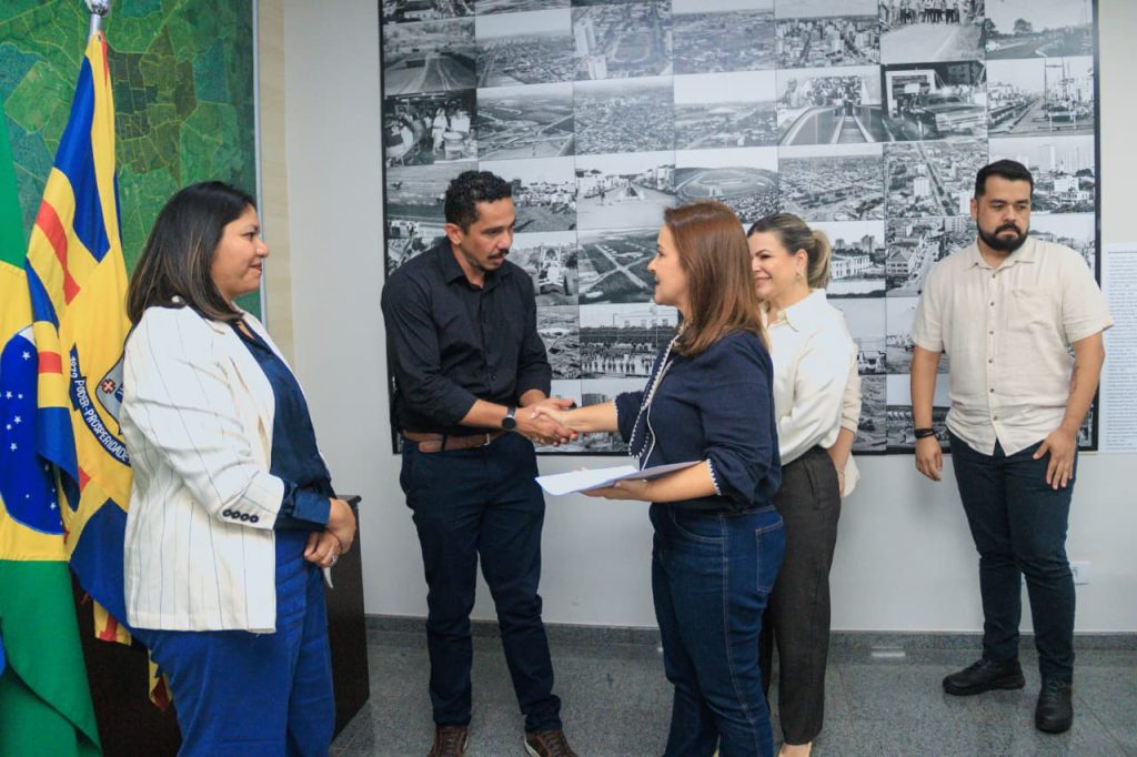 Fotografia de plano inteiro mostrando cinco pessoas de pé em um escritório. No centro, o homem de camisa preta e calça jeans escura aperta a mão da mulher de blusa azul-escura e calça jeans, que segura um papel branco. À esquerda, uma mulher de blazer branco e calça azul observa o aperto de mãos. À direita, outra mulher de blusa branca e calça escura, e um homem de camisa clara e calça escura também acompanham o ato. Ao fundo, há uma parede cinza decorada com um grande painel de fotos em preto e branco e bandeiras oficiais à esquerda.