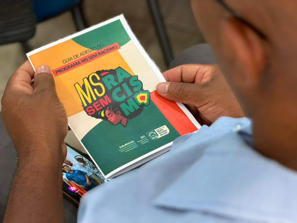 Fotografia em primeiro plano, com foco nas mãos de um homem de pele negra segurando a cartilha "Guia de Adesão do Programa MS sem Racismo". A capa do livreto é dividida em cores verde, laranja e branca, trazendo a ilustração estilizada do perfil de uma mulher negra com cabelo black power e as palavras "MS SEM RACISMO". A foto é tirada por cima do ombro do homem, cujo rosto aparece desfocado em primeiro plano à direita. Ao fundo, um celular repousa sobre seu colo exibindo uma imagem na tela.