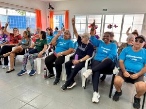 Fotografia colorida em ambiente interno de um grupo de idosos sentados em fileiras de cadeiras brancas e pretas. Vários deles vestem camisetas azuis com a inscrição "CCI Edmundo Scheuneman Piratininga". Uma mulher negra em destaque na frente sorri, enquanto outras pessoas ao seu lado levantam os braços e apontam o indicador para cima, em tom de participação ou resposta. Ao fundo, janelas com cortinas laranjas e outras pessoas assistindo à atividade.