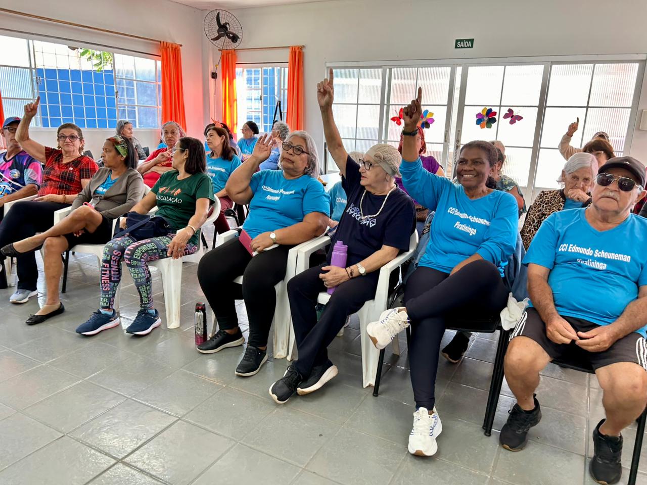 Fotografia colorida em ambiente interno de um grupo de idosos sentados em fileiras de cadeiras brancas e pretas. Vários deles vestem camisetas azuis com a inscrição "CCI Edmundo Scheuneman Piratininga". Uma mulher negra em destaque na frente sorri, enquanto outras pessoas ao seu lado levantam os braços e apontam o indicador para cima, em tom de participação ou resposta. Ao fundo, janelas com cortinas laranjas e outras pessoas assistindo à atividade.