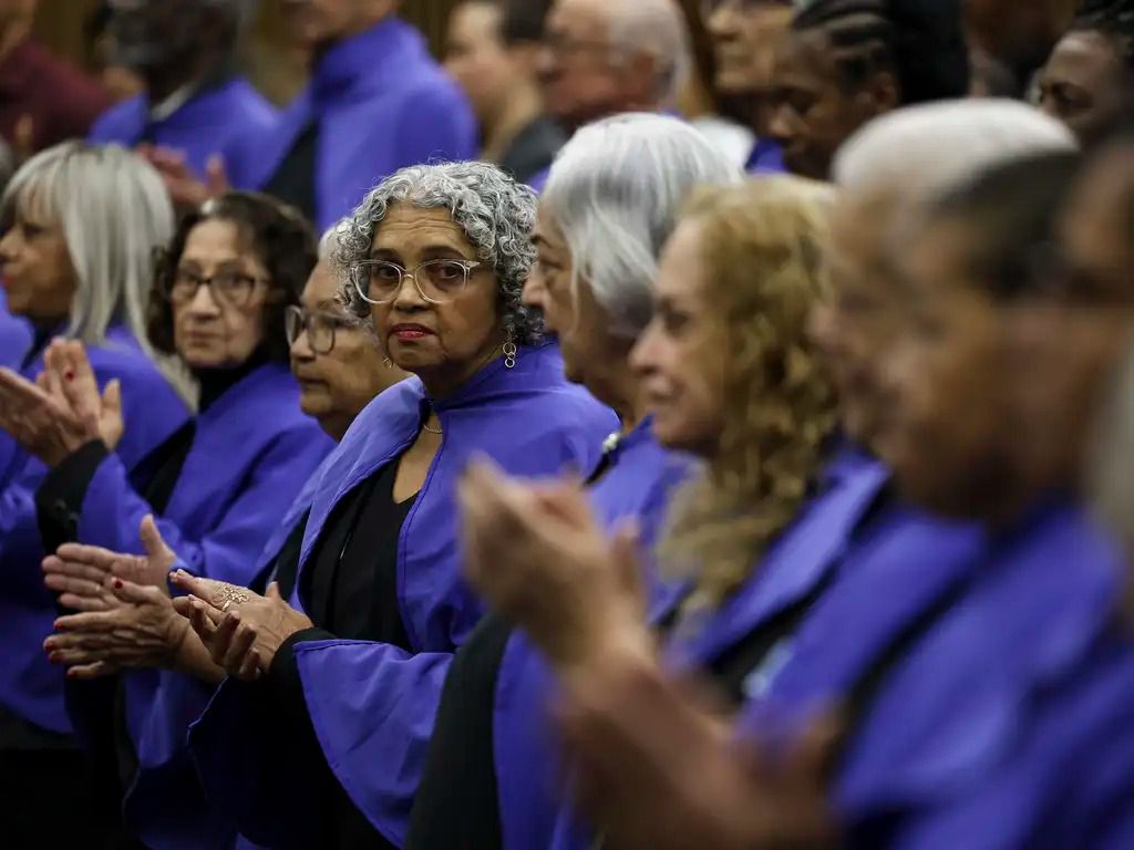 Fotografia colorida com foco seletivo mostrando um grupo de idosos em um coral ou evento formal, todos vestindo batinas roxas. Em destaque na lateral esquerda, uma mulher negra de cabelos grisalhos cacheados e óculos de grau olha em direção à câmera com uma expressão séria. Ao seu redor, outras pessoas aparecem de perfil, com as mãos unidas ou aplaudindo, com o plano de fundo suavemente desfocado.