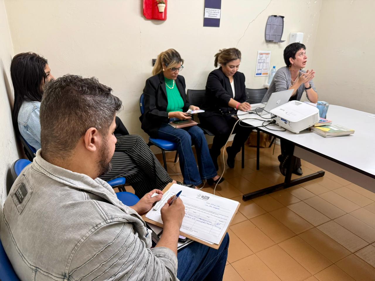Foto em ângulo diagonal mostrando um grupo de pessoas em uma sala de reuniões pequena. Em primeiro plano, à esquerda, um homem de perfil com jaqueta cinza escreve em um papel fixado em uma prancheta. Ao fundo, ao longo de uma mesa branca comprida, três mulheres estão sentadas em cadeiras azuis participando da atividade. Na mesa estão posicionados um notebook, um projetor branco, livros e uma caneca, apontados para a direção de onde as mulheres olham. O piso é de cerâmica clara.