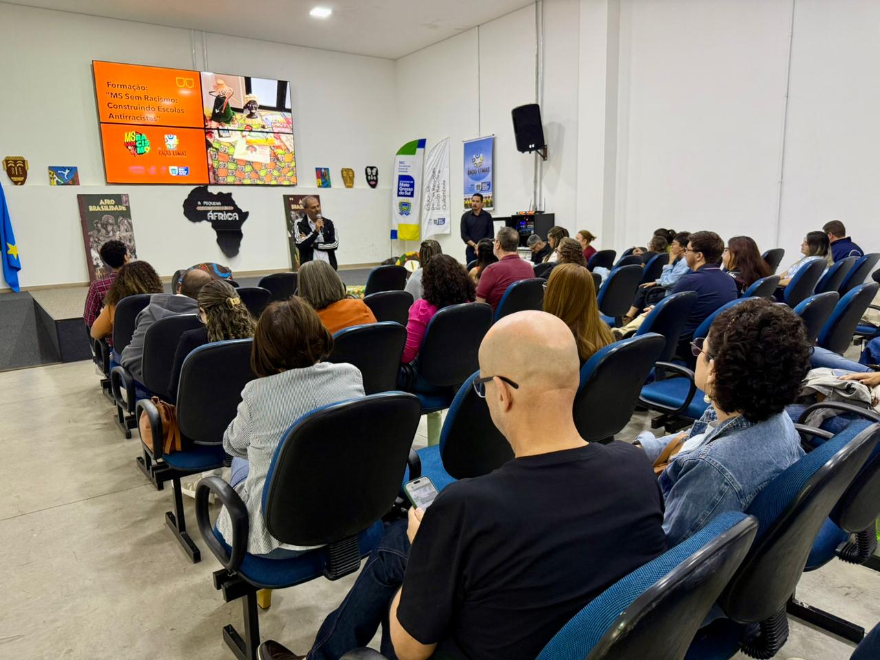 Foto em plano geral mostrando o auditório visto de trás. Várias fileiras de cadeiras azuis estão ocupadas pelo público que assiste atentamente ao evento. No palco ao fundo, um telão quadrado exibe o slide de abertura com o título "Formação: MS Sem Racismo: Construindo Escolas Antirracistas". Próximo ao telão, um homem fala ao microfone.