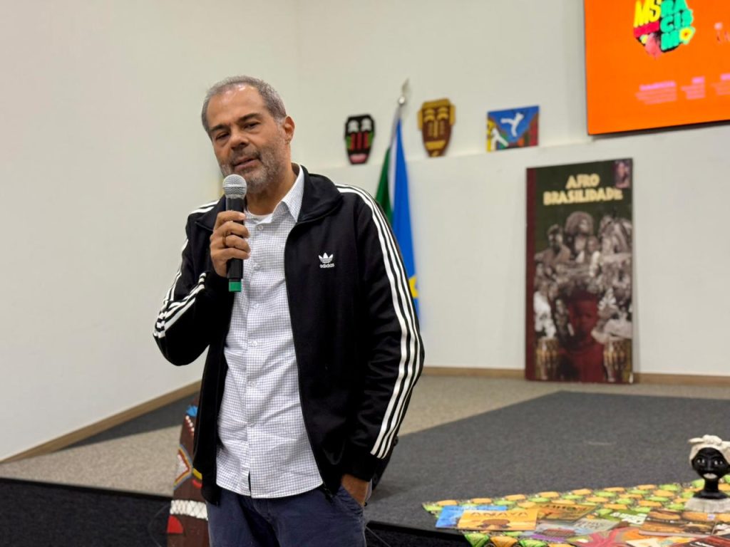 Foto em plano médio centralizada em um homem de pele clara e cabelos grisalhos curtos, usando uma jaqueta esportiva preta com listras brancas nas mangas sobre uma camisa social clara. Ele segura um microfone e fala olhando em direção à câmera. Ao fundo, o palco do auditório exibe decorações na parede e livros dispostos no chão sobre um tecido estampado.