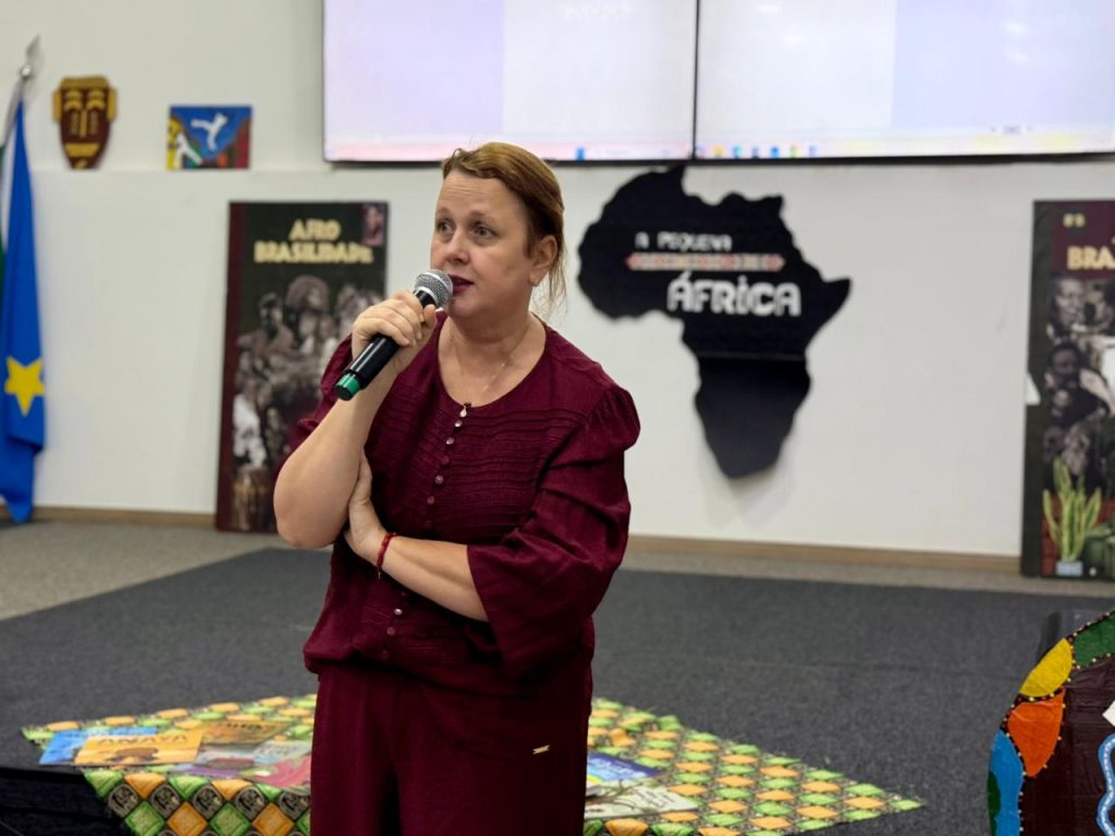 Foto em plano médio focada em uma mulher de pele clara e cabelos castanhos presos, vestindo um conjunto de blusa e calça na cor vinho. Ela segura um microfone com a mão direita e fala olhando para o lado esquerdo da imagem. Ao fundo, destaca-se na parede branca a silhueta preta do mapa da África com a inscrição "A Pequena África".