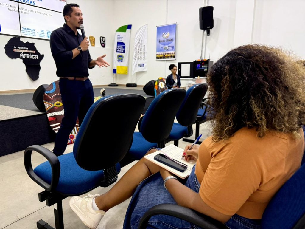 Foto em plano médio no interior de um auditório. No lado esquerdo, no palco, um homem de camisa preta e calça escura fala ao microfone e gesticula. À direita, em primeiro plano e de costas, uma mulher negra de cabelos cacheados e blusa mostarda assiste à palestra sentada em uma cadeira azul, fazendo anotações em um caderno. Ao fundo, veem-se banners institucionais e elementos de decoração afro-brasileira.