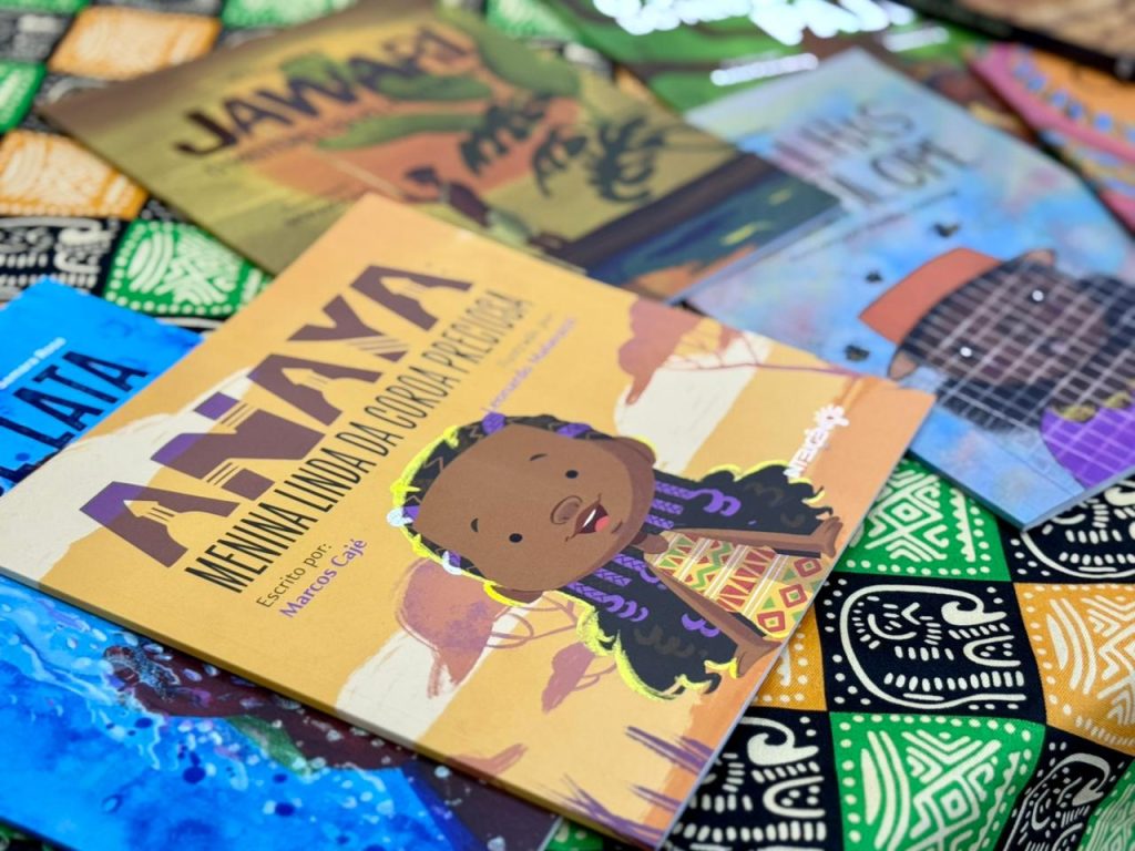Foto em plano detalhe e de ângulo superior mostrando vários livros infantis espalhados sobre um tecido com estampas geométricas afro-indígenas em tons de verde, amarelo e preto. O livro em maior destaque, na parte inferior, tem capa amarela e traz a ilustração de uma menina negra com tranças e trajes coloridos, com o título "Anaya: Menina linda da coroa preciosa", escrito por Marcos Cajé.