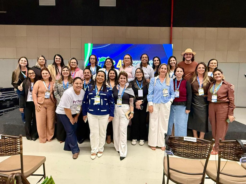 Foto grupal de cerca de vinte e cinco mulheres e um homem posando sorridentes à frente do palco do evento. Eles vestem roupas variadas, muitos usam crachás e estão posicionados em duas fileiras, uma em pé e outra levemente à frente.
