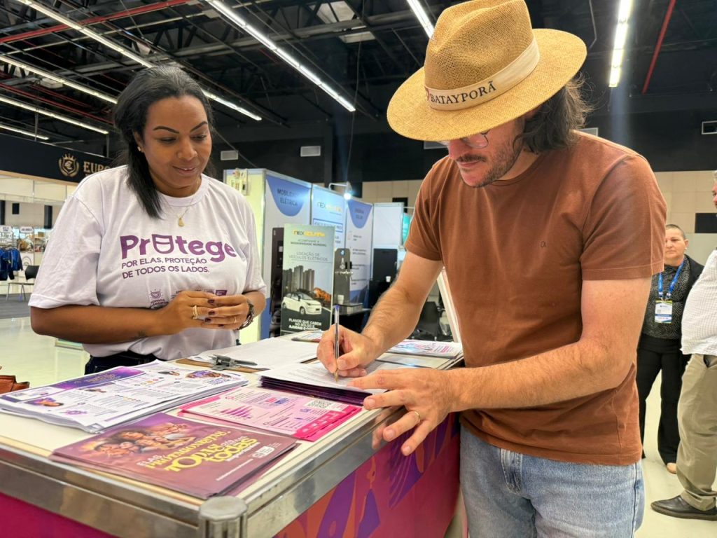 Close de um homem com chapéu de palha escrito "BATAYPORÃ" assinando um documento sobre um balcão. Ao lado dele, uma mulher negra com camiseta branca da campanha "ProTege" observa a assinatura com um sorriso discreto. Há folhetos informativos sobre a mesa.
