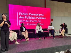 Fotografia de um painel de discussão em um palco. Cinco mulheres estão sentadas em poltronas cinzas, algumas usando fones de ouvido para tradução ou áudio. Ao fundo, um grande telão rosa exibe o texto "Fórum Permanente das Políticas Públicas para Mulheres de Mato Grosso do Sul". No canto esquerdo, em primeiro plano, uma intérprete de Libras faz sinais.