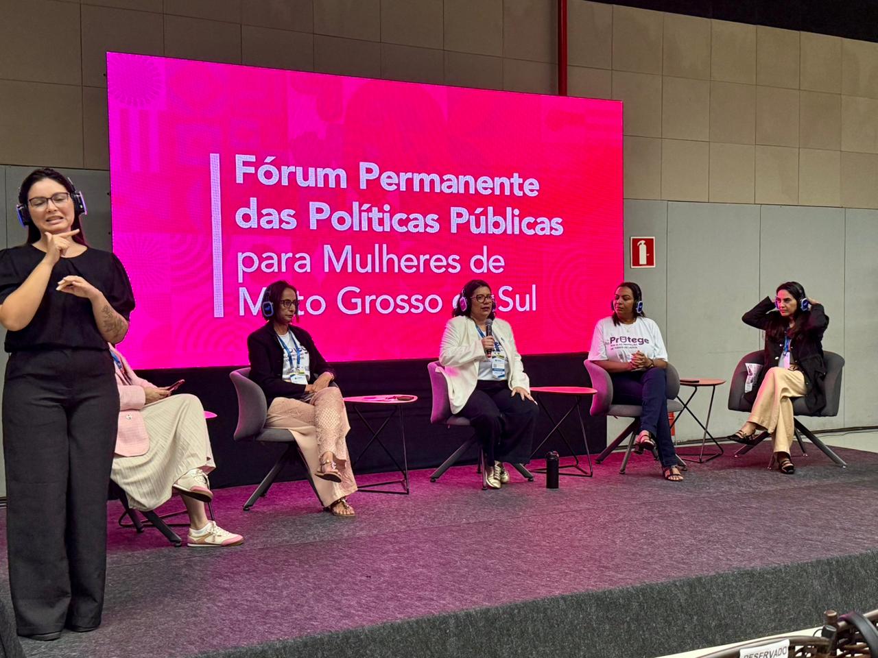 Fotografia de um painel de discussão em um palco. Cinco mulheres estão sentadas em poltronas cinzas, algumas usando fones de ouvido para tradução ou áudio. Ao fundo, um grande telão rosa exibe o texto "Fórum Permanente das Políticas Públicas para Mulheres de Mato Grosso do Sul". No canto esquerdo, em primeiro plano, uma intérprete de Libras faz sinais.