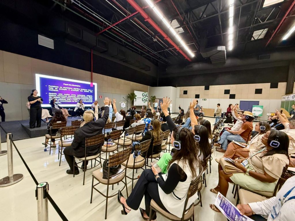 Foto ampla do auditório mostrando o público assistindo ao evento. Muitas pessoas estão sentadas em cadeiras de metal e palha, e várias delas levantam as mãos. No palco ao fundo, a intérprete de Libras e o telão estão visíveis. Quase todos na plateia usam fones de ouvido.