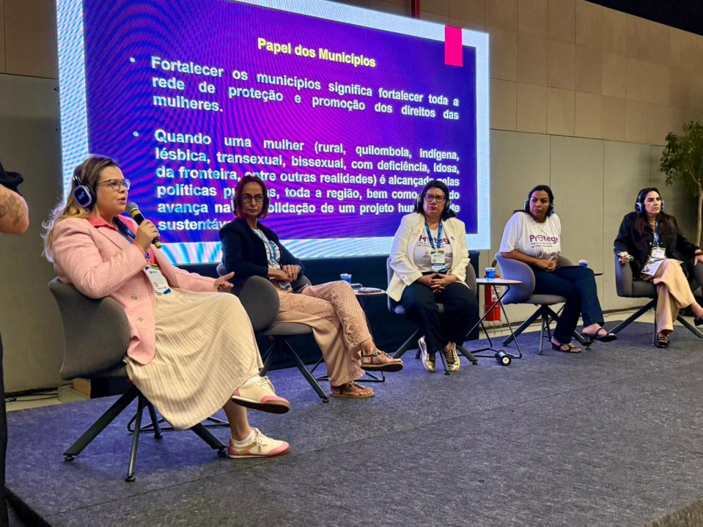 Visão lateral de cinco mulheres sentadas em um painel sobre o palco. A mulher mais à esquerda fala ao microfone. Ao fundo, o telão exibe um slide azul com o título "Papel dos Municípios" e textos sobre o fortalecimento da rede de proteção às mulheres.