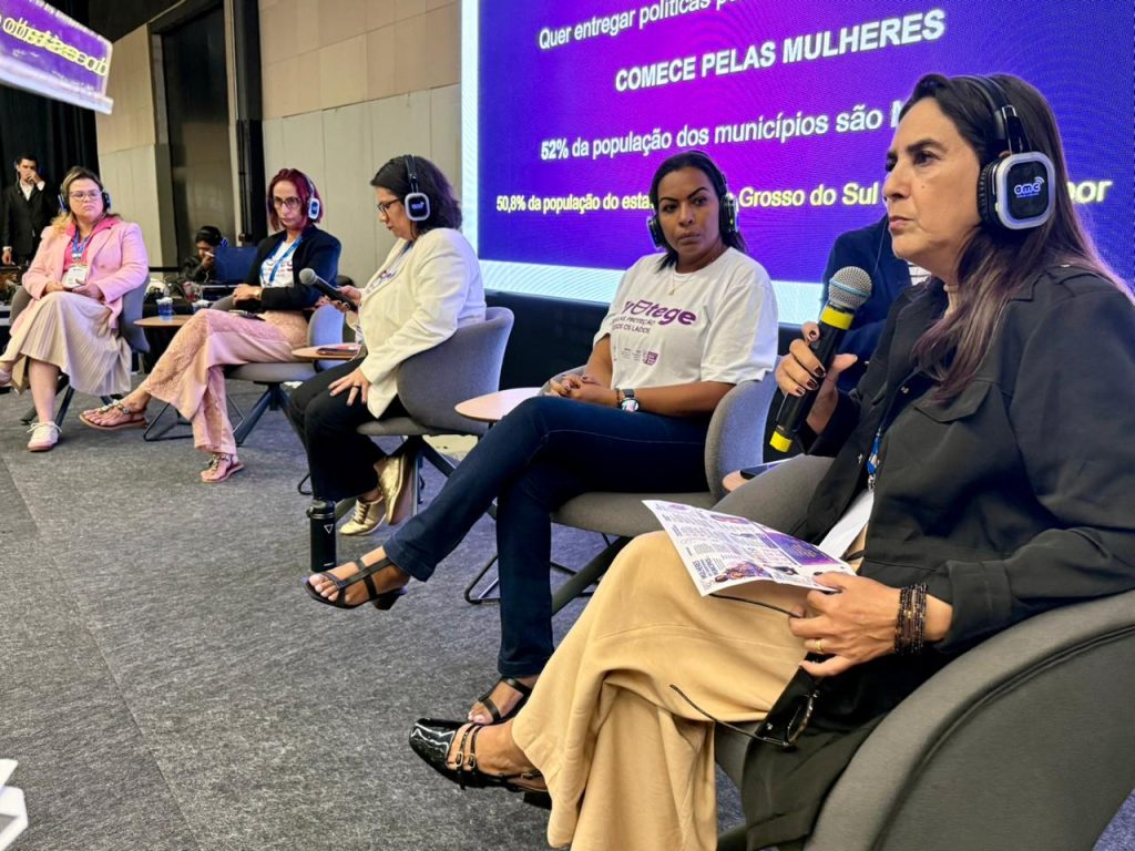 Imagem em ângulo diagonal mostrando quatro mulheres sentadas no painel do palco. A mulher em primeiro plano, à direita, segura um microfone e papéis enquanto escuta. Ao fundo, o telão mostra um slide roxo com a frase em destaque: "COMECE PELAS MULHERES".