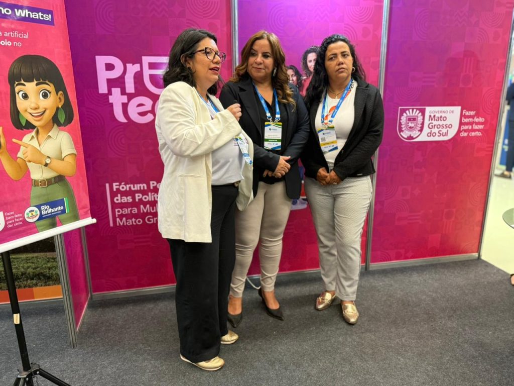 Três mulheres de pé conversando em frente a um painel de fundo rosa. A mulher à esquerda, de blazer branco e óculos, fala ao microfone. As outras duas escutam atentamente. À esquerda, há um banner com a ilustração de uma assistente virtual.