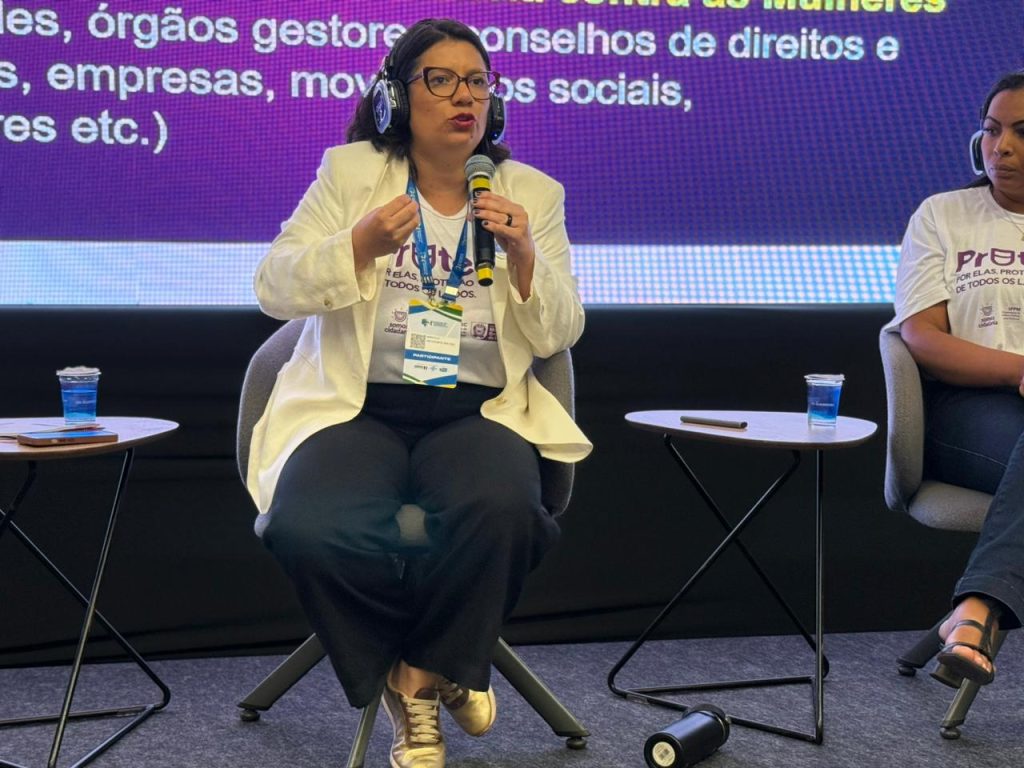 Foto em plano médio focada em uma mulher de óculos e blazer branco sentada no painel, falando ao microfone e gesticulando com a mão direita. Ela usa fones de ouvido grandes e um crachá de participante. Ao lado direito, vê-se parte de outra mulher com camiseta branca escrita "ProTege".