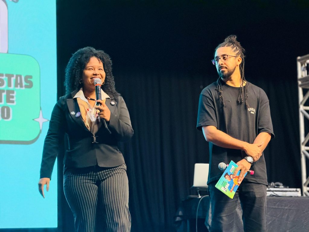 Em um palco com fundo escuro, uma mulher negra com cabelos cacheados volumosos sorri enquanto fala ao microfone, vestindo um terninho preto com calça risca-de-giz. Ao lado dela, à direita, um homem negro com cabelos presos em tranças e óculos, vestindo camiseta preta, segura um folheto colorido do evento "Juventude Plena" e ouve a apresentação.