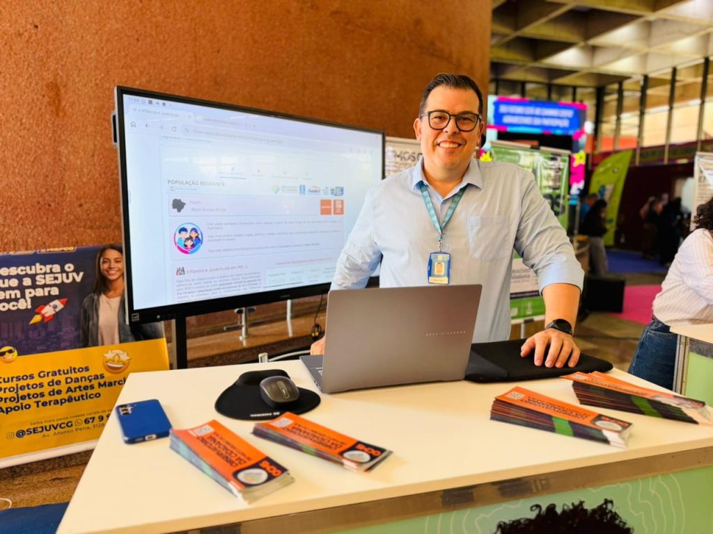 Um homem sorridente de óculos e camisa social azul-clara está de pé atrás de um balcão de atendimento, apoiando a mão em um notebook cinza. Sobre o balcão, há panfletos informativos empilhados, um mouse e um celular azul. Ao fundo, uma tela grande exibe dados do Observatório da Cidadania e o ambiente sugere um pavilhão de eventos institucional.