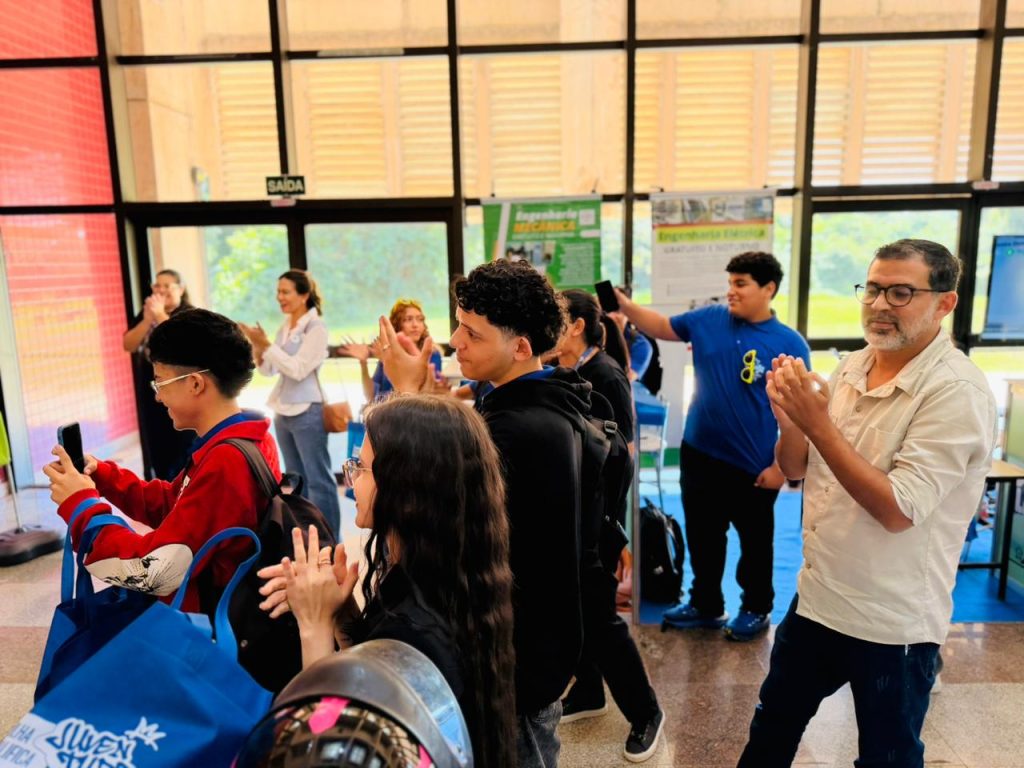 Um grupo de pessoas, incluindo jovens e adultos, aparece reunido em um saguão amplo e iluminado com grandes fachadas de vidro ao fundo. À direita, um homem de óculos e camisa clara bate palmas, acompanhado por outros jovens que também aplaudem ou observam a movimentação. Um dos jovens à esquerda segura um celular para registrar o momento.