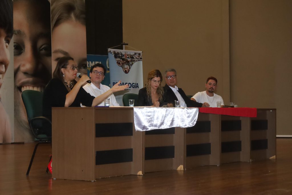 Cinco palestrantes estão sentados atrás de uma longa mesa de madeira no palco de um auditório. Uma mulher à esquerda, de óculos e blusa preta, fala ao microfone gesticulando com o braço esquerdo. Ao lado dela, três homens e outra mulher ouvem atentamente. Uma faixa branca decora o centro da mesa.