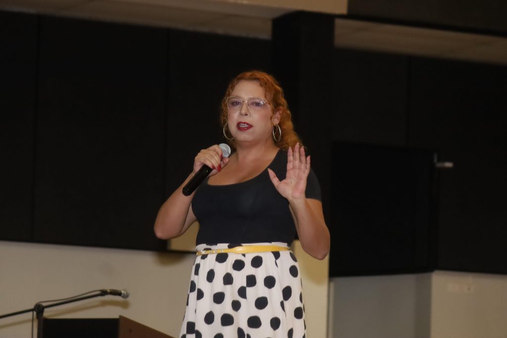 Plano médio de uma mulher com cabelos ondulados acobreados, óculos e argolas douradas, falando ao microfone em um palco escuro. Ela veste uma blusa preta justa e uma saia midi branca com estampa de bolinhas pretas (poá), e está com a mão esquerda levantada.