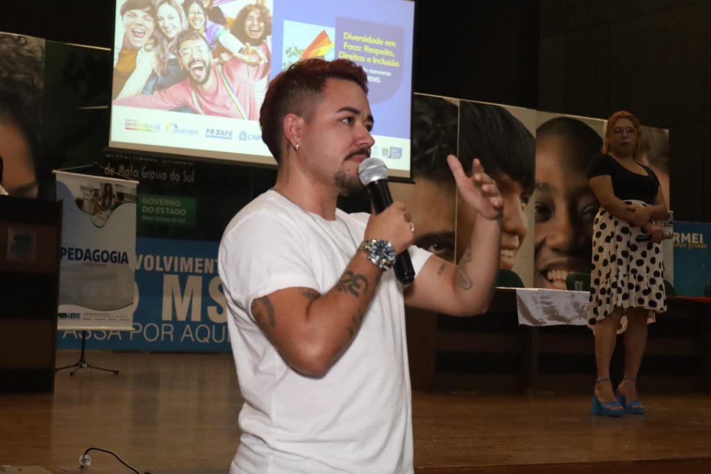 Em foco, de perfil, um homem jovem de cabelo curto avermelhado e barba fala ao microfone gesticulando com o braço esquerdo. Ele veste camiseta branca e tem tatuagens no braço. Ao fundo, no palco, a mulher de saia de bolinhas observa em pé, e o telão do evento aparece iluminado.