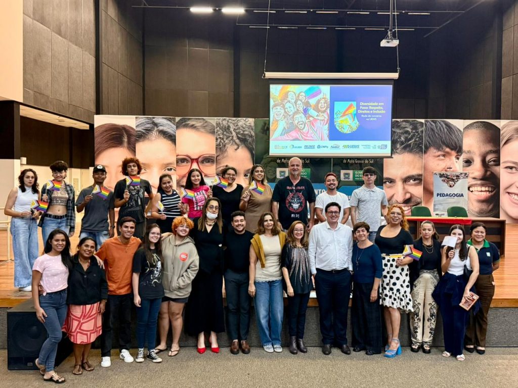 Foto grupal de cerca de 25 pessoas reunidas à frente do palco de um auditório. Algumas pessoas nas pontas e atrás seguram pequenas bandeiras coloridas do orgulho LGBTQIAPN+. Ao fundo, painéis grandes com fotos de rostos diversos e um telão exibem a arte do evento com o título "Diversidade em Foco: Respeito, Direitos e Inclusão na UEMS".
