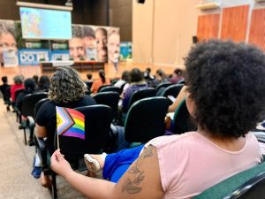 Imagem em perspectiva de dentro de um auditório. Em primeiro plano, à direita, uma pessoa de costas e perfil com cabelo crespo preto e blusa rosa segura uma pequena bandeira do orgulho LGBTQIAPN+ com as cores do arco-íris e o triângulo do progresso. Ao fundo, a plateia sentada em poltronas verdes acompanha uma apresentação no palco, onde há um telão iluminado e painéis com rostos humanos.