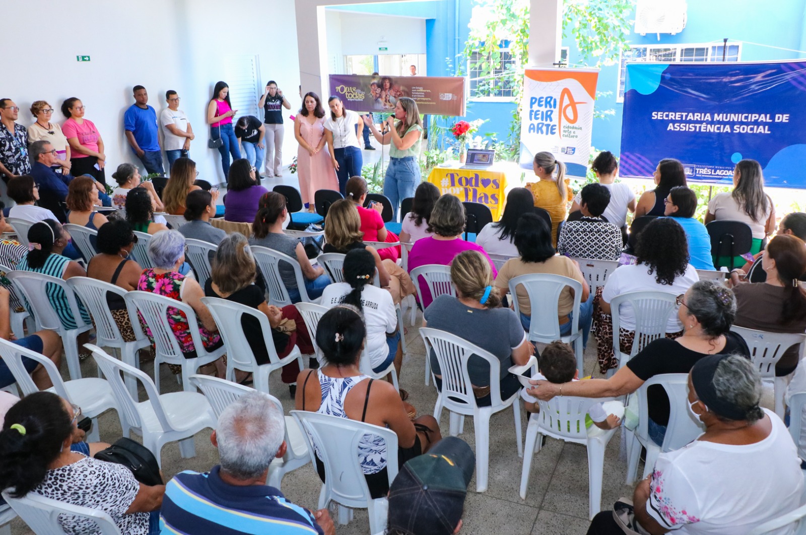 Foto em ambiente iluminado e semiaberto. Em primeiro plano, uma plateia majoritariamente feminina está sentada em cadeiras de plástico brancas, de costas para a câmera. Ao fundo, uma mulher em pé fala ao microfone. Atrás dela, há um banner com a identidade visual do projeto "Perifeiarte" e outras pessoas em pé acompanhando o evento.
