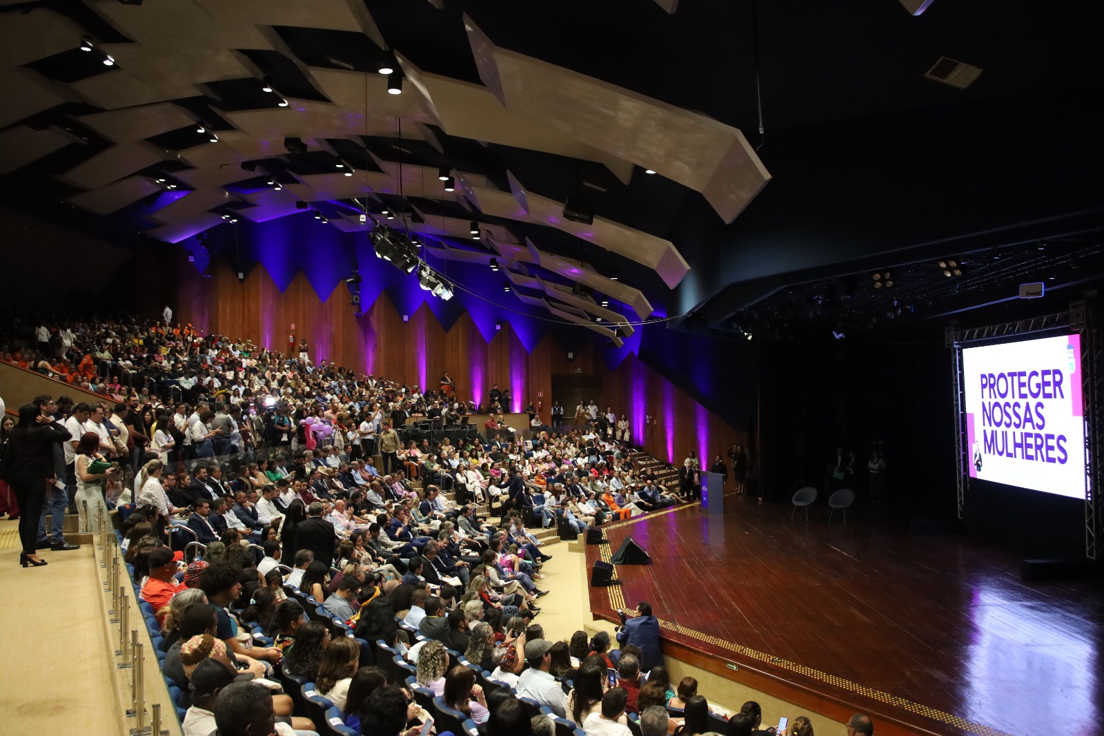 Fotografia em plano geral e perspectiva diagonal de um grande auditório lotado durante um evento. À esquerda, a plateia ocupa as poltronas dispostas em arquibancadas inclinadas. À direita, há um palco espaçoso de madeira escura com um grande telão iluminado que exibe a frase "PROTEGER NOSSAS MULHERES" em letras roxas sobre um fundo branco. O teto do auditório possui painéis geométricos modernos e a iluminação cênica destaca as paredes laterais com tons de roxo.