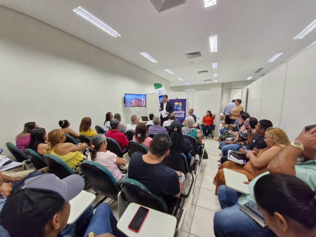 Foto em ângulo aberto, tirada de trás, mostrando uma plateia sentada em cadeiras escuras com pranchetas acopladas. As pessoas assistem a um homem de paletó escuro que fala em pé, à frente da sala, ao lado de um banner roxo onde se lê "Receita Cidadã". No fundo, há uma tela de TV ligada e divisórias brancas de escritório.
