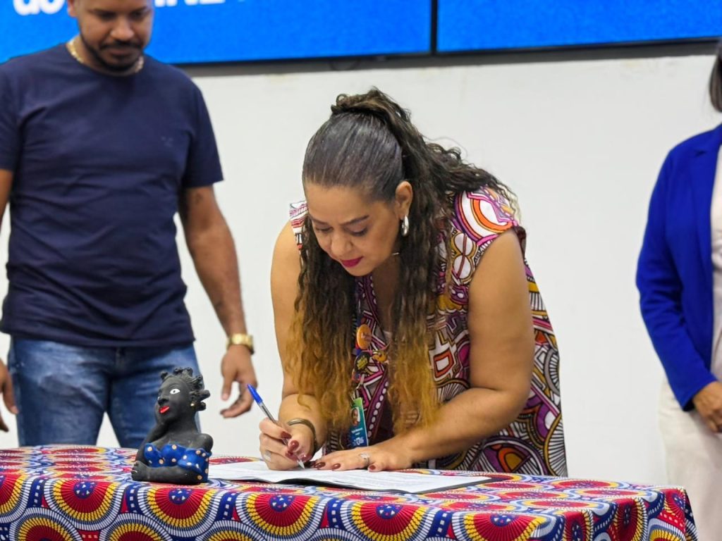 Uma mulher com cabelos longos e ondulados presos em um rabo de cavalo aparece inclinada, assinando um documento sobre uma mesa coberta com uma toalha de estampa étnica colorida. Ela veste uma blusa estampada em tons de rosa, roxo e amarelo. Uma pequena escultura de cerâmica preta com detalhes azuis decora a mesa, enquanto outras pessoas aparecem ao fundo.