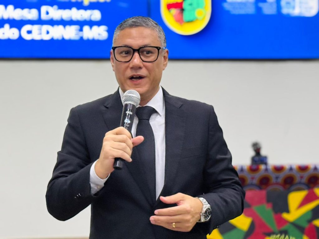 Close-up de um homem de meia-idade, com cabelos grisalhos curtos e óculos de armação preta grossa, falando ao microfone. Ele veste um terno preto clássico, camisa branca e gravata preta. O fundo está ligeiramente desfocado, mostrando parte do telão azul do evento.