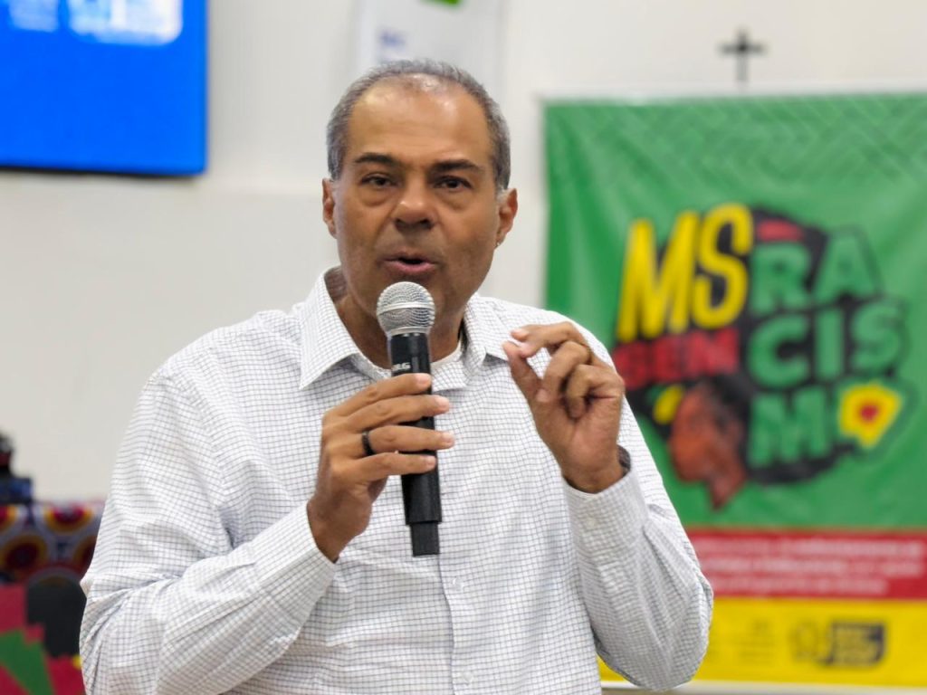 Fotografia em plano médio de um homem de pele parda e cabelos curtos grisalhos, vestindo uma camisa social branca com um padrão xadrez bem fino. Ele está de pé, falando ao microfone que segura com a mão direita, enquanto gesticula com a mão esquerda. Ao fundo, de forma desfocada, há uma bandeira verde com letras coloridas onde se lê "MS" em amarelo e "RACISMO" em verde e vermelho.