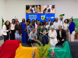 Foto oficial de grupo com cerca de vinte participantes reunidos no palco do evento. Homens e mulheres de diversas idades sorriem para a foto; alguns estão em pé e outros sentados na primeira fileira, incluindo um homem em cadeira de rodas e outro com vestes tradicionais brancas e torço na cabeça. À frente do grupo, há tecidos decorativos nas cores vermelha, amarela e verde, além de arranjos de plantas. O telão azul serve como plano de fundo.