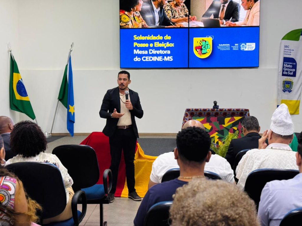 Vista do público de um auditório assistindo a um homem jovem, de pele parda, cabelo curto e bigode, que fala em pé ao centro segurando um microfone. Ele veste paletó preto sobre uma camiseta bege e calça preta. Ao fundo, há um grande telão azul com o texto "Solenidade de Posse e Eleição: Mesa Diretora do CEDINE-MS" e as bandeiras do Brasil e de Mato Grosso do Sul nas laterais.